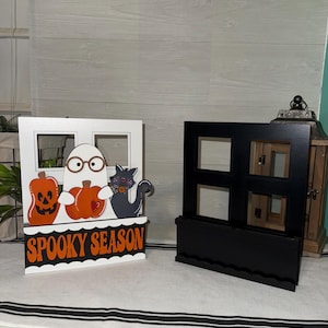 May include: Two Halloween-themed wooden decorations. One features a ghost with glasses, pumpkins, and a black cat, with the text "Spooky Season." The other is a black, window-shaped display shelf.