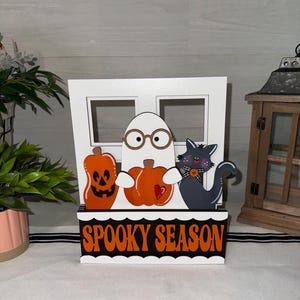 Peut inclure: Décoration en bois sur le thème d'Halloween avec un fantôme blanc portant des lunettes, deux citrouilles oranges et un chat noir. La base indique "Spooky Season" en lettres oranges. La décoration est sur fond blanc.