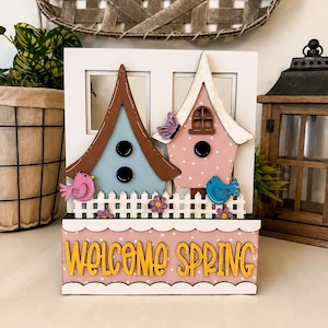 Puede incluir: Un letrero decorativo de madera con temática primaveral con dos casas para pájaros, una valla y la inscripción "WELCOME SPRING". Las casas para pájaros son azules y rosas con tejados marrones. Un pájaro rosa y otro azul están posados en la valla.