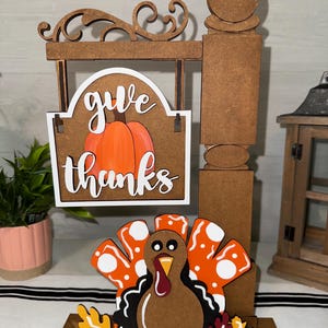 Könnte beinhalten: Holz-Dekoration zum Erntedankfest mit einem Schild mit der Aufschrift "give thanks" und einer Kürbis-Grafik. Eine Truthahn mit orangefarbenen und weißen Federn und Herbstblättern befindet sich am Sockel. Das Schild und der Truthahn sind braun, orange und weiß.