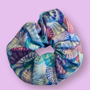 May include: A fabric scrunchie with a seashell pattern in shades of blue, purple, and pink. The scrunchie is a hair accessory, perfect for adding a touch of style to any hairstyle. The design features detailed illustrations of seashells.