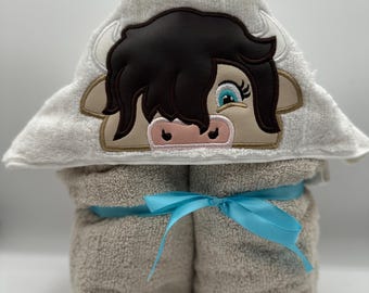 May include: A beige hooded baby towel featuring an embroidered cow face. The cow has brown hair, tan ears, and blue eyes. The towel is tied with a light blue ribbon.