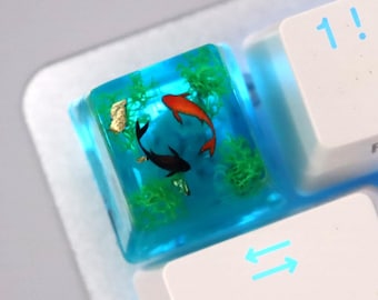 May include: A white keyboard with a blue backlight. The '1' key has been replaced with a custom keycap featuring a blue resin cube with two goldfish swimming inside.