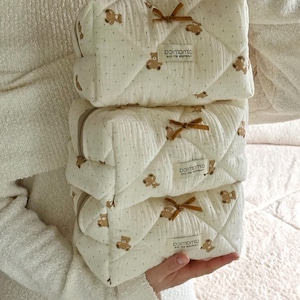 Könnte beinhalten: Drei weiße gesteppte Kosmetiktaschen mit braunen Teddybär-Designs und schwarzen Punkten. Jede Tasche hat eine braune Schleife und ein Etikett mit der Aufschrift "paimomio and the experience".