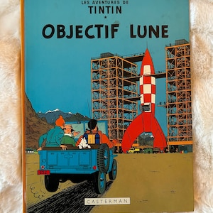 Puede incluir: Una portada de cómic vintage que muestra un jeep azul alejándose de una gran plataforma de lanzamiento de cohetes. La portada se titula "Objectif Lune" y presenta el texto "Les Aventures de Tintin" y "Herge".