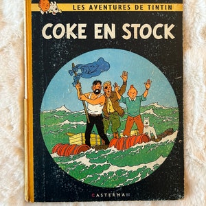 May include: A vintage comic book cover featuring Tintin, a Belgian reporter, and his dog Snowy. The cover is black with a yellow border and the title "COKE EN STOCK" in red letters. The image shows Tintin and his friends on a raft in the middle of the sea.