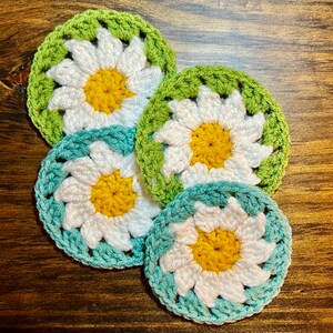 May include: Four crocheted daisy coasters in shades of green and blue. The coasters feature a white daisy with a yellow center.