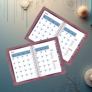 Könnte beinhalten: Zwei offene Spiralplaner mit einem Januar-Kalenderlayout. Die Seiten sind weiß mit blauen Rasterlinien und blauen Punktmarkierungen. Die Einbände sind ein gedämpftes Rot-Violett mit einem genähten Rand.