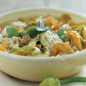 Puede incluir: Primer plano de un cuenco lleno de sopa de verduras. La sopa contiene rodajas de calabacín, flores de calabaza naranja y granos. Hojas de menta fresca adornan la sopa. El cuenco es de color crema claro.