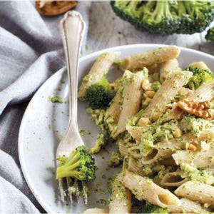 Puede incluir: Pasta penne con brócoli y pesto, adornada con nueces, en un plato blanco. Un tenedor plateado sostiene un trozo de brócoli. El fondo presenta un paño gris y un ramillete de brócoli.