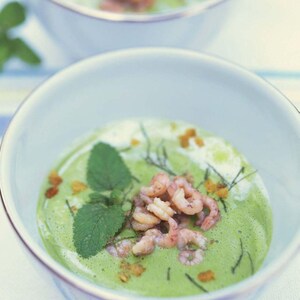 Puede incluir: Primer plano de dos cuencos blancos llenos de sopa verde, adornados con camarones, hojas de menta y pequeñas flores amarillas. La sopa tiene una textura cremosa y es de color verde vibrante.