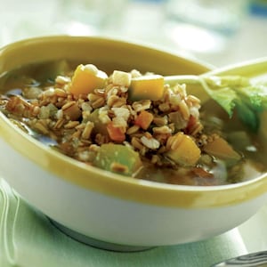 Puede incluir: Primer plano de un cuenco de sopa con borde amarillo. La sopa contiene granos visibles, verduras cortadas en cubitos y caldo. Las verduras incluyen trozos naranjas, verdes y blancos. Una hoja verde es visible en el lado derecho del cuenco.