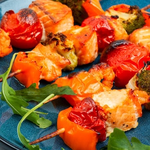 Puede incluir: Primer plano de un plato azul con brochetas a la parrilla. Las brochetas incluyen trozos de salmón, pimientos naranjas, tomates rojos y brócoli. Hojas frescas de rúcula adornan las brochetas, creando una presentación colorida y apetitosa.