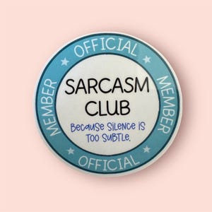 May include: A round, white button with a light blue border and text that reads "OFFICIAL MEMBER SARCASM CLUB" and "Because Silence is Too Subtle." The button has a light pink background.