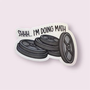 May include: A sticker featuring a stack of gray weight plates with the text "SHHH... I'M DOING MATH" in black. The sticker has a white border and is set against a light pink background.