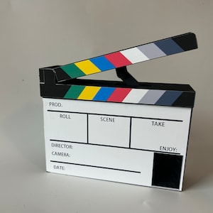 May include: A black and white clapperboard with colorful stripes. The clapperboard has labels for "Prod.", "Roll", "Scene", "Take", "Director", "Camera", "Date", and "Enjoy".