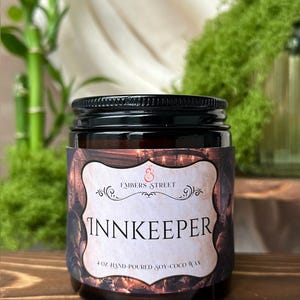 May include: A 4 oz amber glass jar candle with a black screw-top lid. The label features a vintage-style illustration of a tavern scene with the text Embers Street, INNKEEPER, and 4 oz hand-poured soy-coco wax printed in black and red ink.