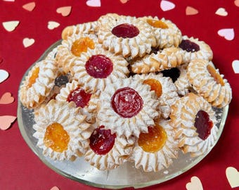 May include: A pile of round, scalloped-edge shortbread cookies dusted with powdered sugar, each featuring a central dollop of either bright red or orange fruit jam, arranged on a clear glass plate against a deep red background.