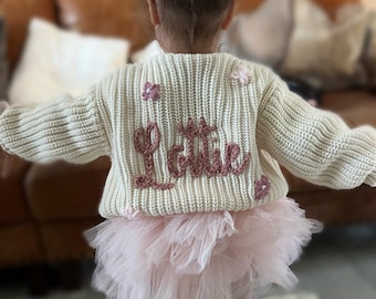 Può includere: Una bambina indossa un maglione di lana color crema con il nome "Lottie" ricamato in filo rosa. Il maglione è decorato con fiori rosa. Indossa anche una gonna di tulle rosa.