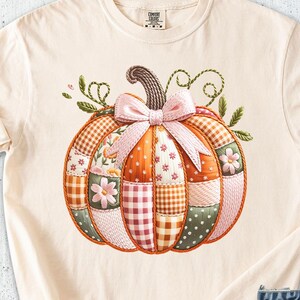 May include: Ivory-colored t-shirt featuring a colorful patchwork pumpkin design with a pink bow. The shirt is paired with denim shorts, a brown hat, and cream-colored shoes. A notebook, sunglasses, and pen are also visible.