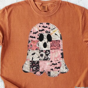 May include: An orange t-shirt featuring a patchwork ghost design with black, pink, and white patterns. The shirt is styled with denim shorts, a brown hat, and cream-colored shoes. A notebook, pen, and sunglasses are also visible.
