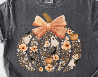 May include: Dark gray t-shirt with a pumpkin design. The pumpkin is decorated with flowers, ghosts, and a bow. The word "PEPPER" is printed at the top. The shirt is paired with denim shorts, shoes, and a hat.