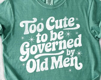 May include: A light green t-shirt with the text "Too Cute to be Governed by Old Men" in white. The image also includes denim shorts, a brown hat, sunglasses, a notebook, shoes, and flowers.