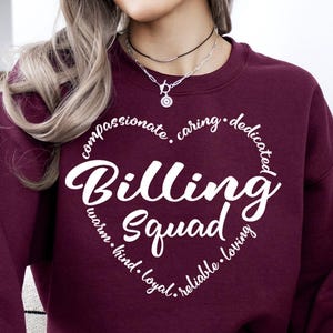 May include: A maroon sweatshirt with the words "Billing Squad" in a heart-shaped design. The heart includes words like "compassionate," "caring," and "loving." The sweatshirt is a solid color and has a crew neck.