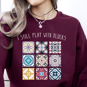 May include: A maroon sweatshirt with the text "I STILL PLAY WITH BLOCKS" above a grid of colorful, patterned tile designs. The tiles feature various geometric and floral patterns in shades of blue, yellow, red, and teal.
