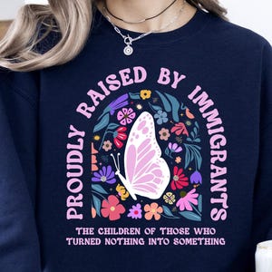 A woman wearing a navy blue sweatshirt with a colorful graphic design and text, sitting on a striped blanket and holding a white mug.