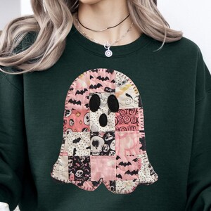 May include: Dark green sweatshirt featuring a patchwork ghost design. The ghost is composed of various fabric squares in pink, black, and white, with skull and bat patterns. The sweatshirt has a crew neck and long sleeves, perfect for Halloween.