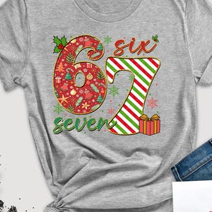 May include: A gray t-shirt with the numbers "6" and "7" in a Christmas theme. The number "6" is red with Christmas icons, and the "7" is red and green striped. The word "six" is above the "7" and the word "seven" is below.