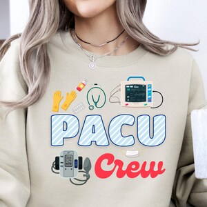 May include: Beige sweatshirt with the text "PACU Crew" in blue and red, featuring medical equipment graphics. The design includes a stethoscope, gloves, and a heart rate monitor. A white mug is held in the hand.