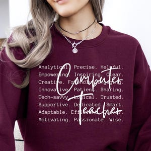 May include: A maroon sweatshirt with the word "Computer" and "Teacher" in white script. The sweatshirt has a list of descriptive words such as "Analytical", "Inspiring", and "Patient". The model is wearing a necklace and white sweatpants.