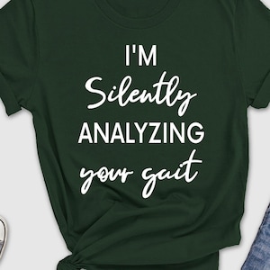 May include: Dark green t-shirt with the text "I'M Silently ANALYZING your gait" in white. The shirt is styled with a pair of blue jeans, sunglasses, sneakers, a hat, and a small potted plant.