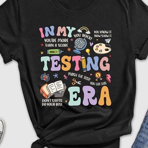 May include: Black t-shirt with a colorful graphic design that says "In My Testing Era". The graphic includes a rainbow, a pencil, a paint palette, a book, and other school-related items.