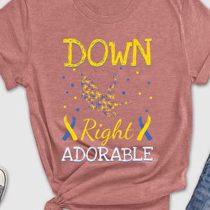 May include: A dusty rose t-shirt with the words "DOWN Right ADORABLE" in yellow and white, with blue and yellow accents. The shirt is styled with blue jeans, sunglasses, and sneakers.
