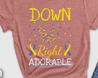 May include: A dusty rose t-shirt with the words "DOWN Right ADORABLE" in yellow and white, with blue and yellow accents. The shirt is styled with blue jeans, sunglasses, and sneakers.