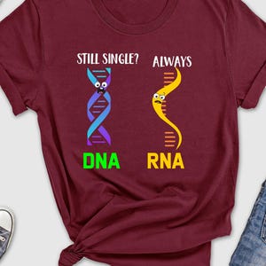 May include: Burgundy t-shirt with a science-themed graphic. The graphic shows two DNA strands, one labeled "DNA" and the other labeled "RNA". The text "Still single? Always" is above the graphic.
