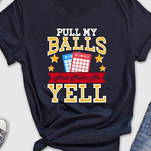May include: Navy blue t-shirt with the text "PULL MY BALLS And Make Me YELL" in red and yellow. The text is surrounded by red stars and a bingo card graphic.