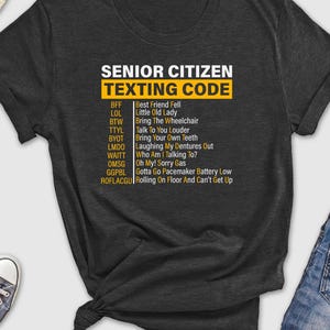 May include: A dark gray t-shirt with a white graphic that says "Senior Citizen Texting Code" and lists common texting abbreviations and their meanings.
