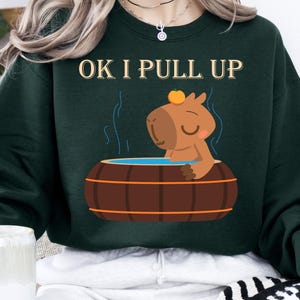 May include: Dark green sweatshirt with the text "OK I PULL UP" above a cartoon capybara relaxing in a wooden hot tub. The capybara is brown with a small orange fruit on its head. The sweatshirt is a crew neck style.