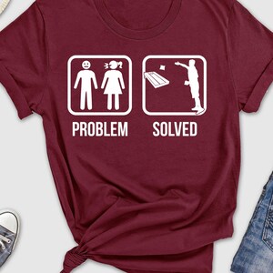 May include: Burgundy t-shirt with a white graphic of a stick figure couple with the text "Problem" and a stick figure throwing a beanbag with the text "Solved".