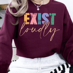 May include: A maroon cropped sweatshirt with the text "EXIST LOUDLY" in colorful letters. The letters are in a distressed font and have a rainbow color scheme.