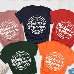 May include: A flat lay featuring six t-shirts in various colors: heather mauve, navy, red, forest green, and orange. Each shirt has a white circular graphic with the text "Making a Difference" and "Neuro Squad". Accessories include a straw hat, sunglasses, and denim jeans.