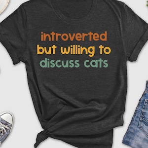 May include: A dark gray t-shirt with the text "introverted but willing to discuss cats" in a retro font with orange, green, and yellow colors.