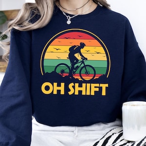 May include: Navy blue sweatshirt with a retro sunset graphic featuring a person riding a bicycle. The text "OH SHIFT" is printed below the graphic.