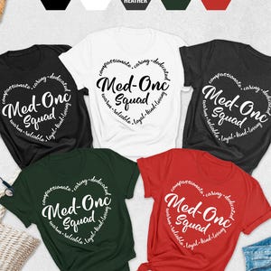 May include: A collection of crew neck t-shirts in black, white, dark grey heather, forest green, and red. Each shirt features the text "Med-One Squad" inside a heart-shaped design, surrounded by words like "compassionate" and "caring".