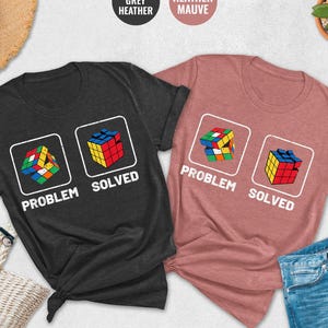 May include: Two t-shirts with a Rubik's cube graphic and the text "Problem Solved". The shirt on the left is dark heather grey and the shirt on the right is heather mauve.