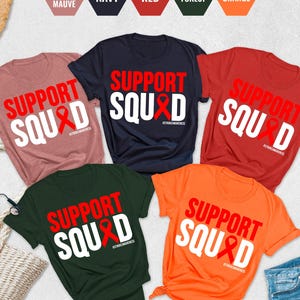 May include: Five t-shirts in different colors with the text "Support Squad" and a red ribbon printed on them. The colors of the shirts are heather mauve, navy, red, forest green, and orange.